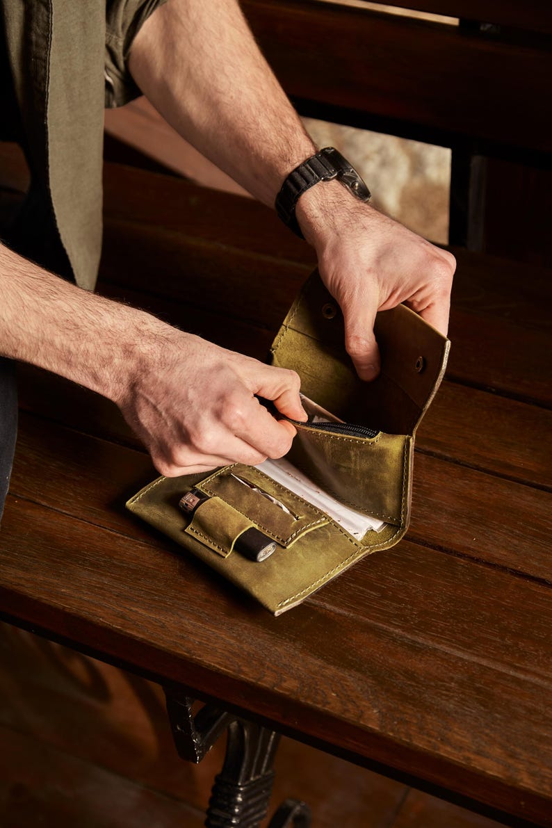 May include: An olive green leather wallet featuring a fold-over design with a snap button closure. The interior includes a zippered compartment, a dedicated slot for a small cylindrical tool, and an open pocket containing white paper documents.