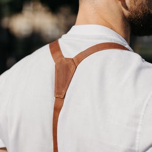 May include: A person wearing a white shirt with brown leather suspenders. The suspenders are attached to the shirt with a leather patch at the back. The leather is a light brown colour. The background is blurred.