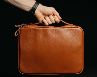 May include: A rectangular, brown leather briefcase with a top handle and a zipper closure. The briefcase is held by a person's hand against a black background. The leather has a textured appearance.
