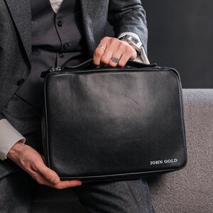 May include: A black leather briefcase with the text "JOHN GOLD" in white lettering. The briefcase is rectangular with a zipper closure. The image shows a person wearing a grey suit and a white shirt.