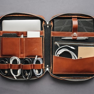 May include: Open, brown leather organizer with multiple compartments. Contains a laptop, phone, notebook, pen, cables, and a small notepad. The organizer is zippered and has a black interior with brown leather accents.