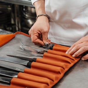 Könnte beinhalten: Eine rostfarbene Messertasche aus Leder mit mehreren Innenfächern, die verschiedene Küchenmesser aus Edelstahl, kulinarische Pinzetten und eine Gabel enthalten, mit grauem Innenfutter und einem sicheren Metallverschluss.