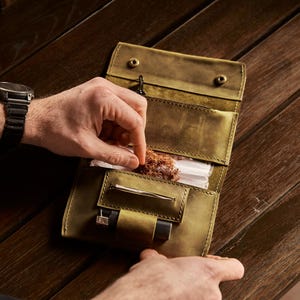 May include: An olive green leather tobacco pouch featuring a fold-over design with two metal snap closures. The interior includes a dedicated compartment for loose tobacco, a horizontal slot for rolling papers, and a small loop holding a black lighter.
