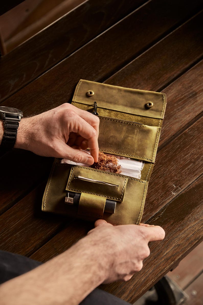May include: An olive green leather tobacco pouch featuring a fold-over design with two metal snap closures. The interior includes a dedicated compartment for loose tobacco, a horizontal slot for rolling papers, and a small loop holding a black lighter.