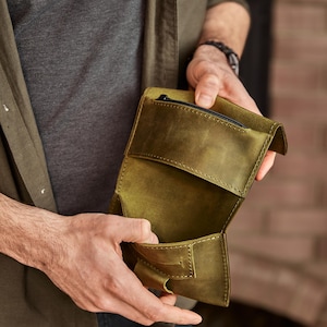 May include: An olive green leather wallet featuring a fold-over design with visible stitching along the edges. The wallet includes an interior compartment and a small zippered pocket at the top, showcasing a matte texture and a structured build.