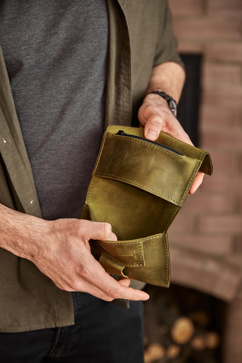 May include: An olive green leather wallet featuring a fold-over design with visible stitching along the edges. The wallet includes an interior compartment and a small zippered pocket at the top, showcasing a matte texture and a structured build.