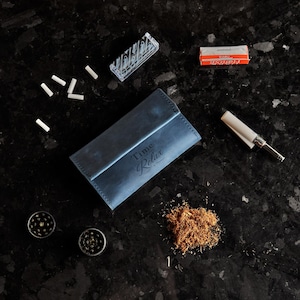 May include: A blue leather tobacco pouch with the text Time to Relax embossed on the front, accompanied by a small pile of loose tobacco, two metal grinders, a pack of rolling papers, a box of filter tips, and a cigarette rolling machine.