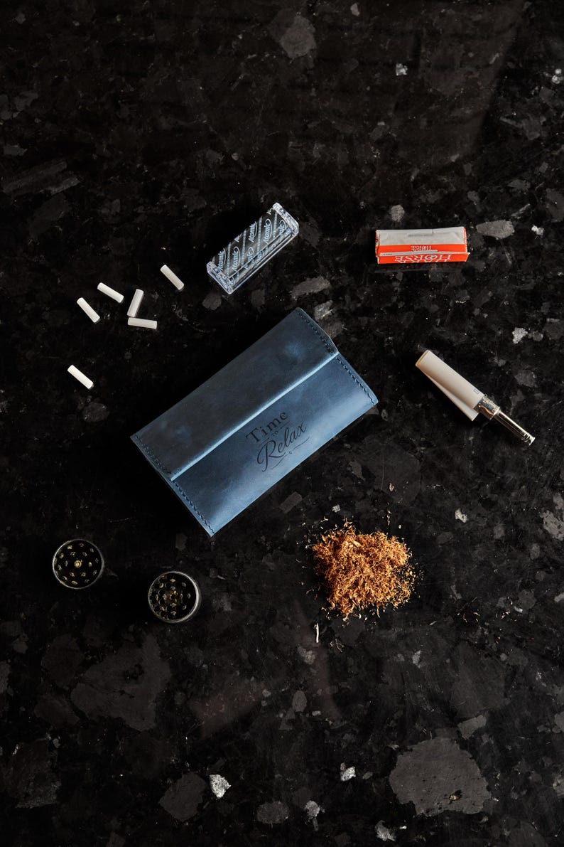 May include: A blue leather tobacco pouch with the text Time to Relax embossed on the front, accompanied by a small pile of loose tobacco, two metal grinders, a pack of rolling papers, a box of filter tips, and a cigarette rolling machine.