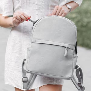 May include: A light gray backpack with a rounded top and a textured surface. The backpack has adjustable straps, a top handle, and a front pocket. The zipper pulls are silver. The bag is held by a person wearing a white dress.