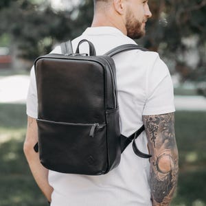 Puede incluir: Una mochila de cuero negro con forma rectangular y un bolsillo frontal. La mochila tiene cierre de cremallera y correas ajustables. La mochila la lleva una persona con una camisa blanca.