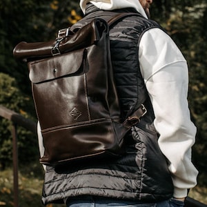 May include: A dark brown leather roll-top backpack featuring a front flap pocket with a snap closure, a metal buckle strap at the top, and a small embossed logo on the lower front section. The bag has adjustable leather shoulder straps and side buckles.