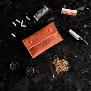 May include: A tan leather tobacco pouch with the initials H.J.P. embossed on the front flap. The pouch is surrounded by loose tobacco, two small metal grinders, a pack of rolling papers, a clear box of filter tips, and a cigarette rolling machine.