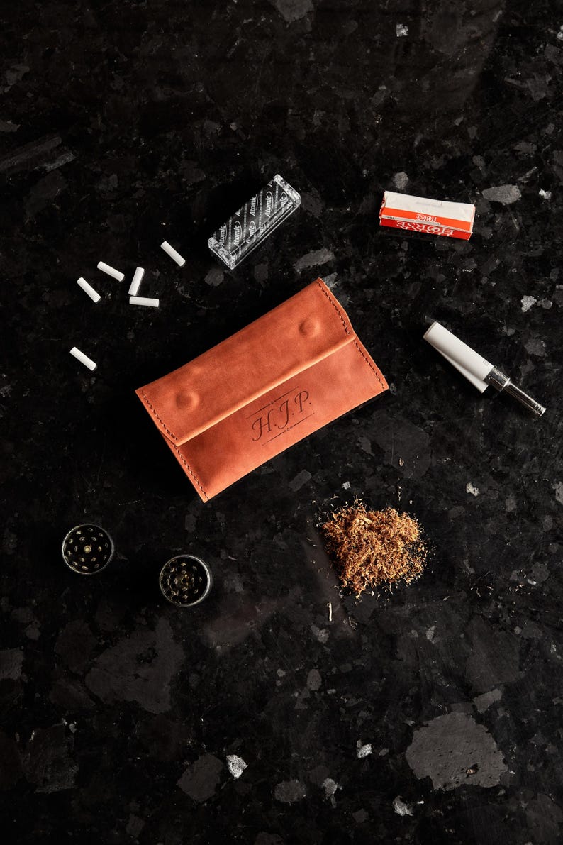 May include: A tan leather tobacco pouch with the initials H.J.P. embossed on the front flap. The pouch is surrounded by loose tobacco, two small metal grinders, a pack of rolling papers, a clear box of filter tips, and a cigarette rolling machine.