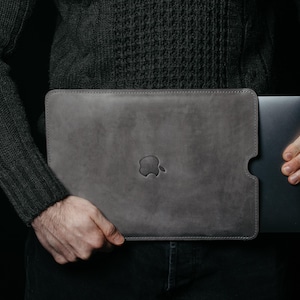 Könnte beinhalten: Eine graue Laptop-Hülle aus Leder mit dem geprägten Apple-Logo auf der Vorderseite. Die Hülle wird von einer Person gehalten, die einen dunkelgrauen Pullover und eine schwarze Hose trägt. Der Laptop ist teilweise sichtbar.