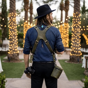May include: An olive green leather camera harness with an X-shaped back design, metal hardware, and adjustable straps. The harness supports a camera on the right side and a small matching leather pouch on the left side, worn over a dark blue shirt.