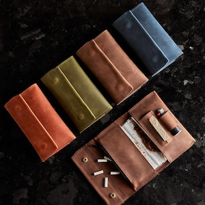 May include: Four rectangular leather tobacco pouches in orange, olive green, brown, and blue, arranged diagonally. One brown pouch is open, revealing a clear plastic tobacco pouch, a small lighter, and several white cigarette filters inside.