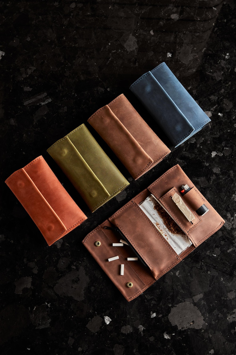 May include: Four rectangular leather tobacco pouches in orange, olive green, brown, and blue, arranged diagonally. One brown pouch is open, revealing a clear plastic tobacco pouch, a small lighter, and several white cigarette filters inside.