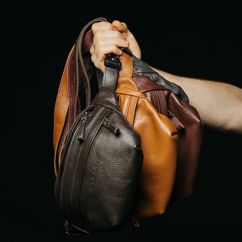 Puede incluir: Una mano sostiene varias bandoleras de cuero en diferentes colores: marr&oacute;n oscuro, marr&oacute;n claro, burdeos y gris. Cada bolso tiene un compartimento principal con cremallera y una correa ajustable. Los bolsos son de cuero texturizado.
