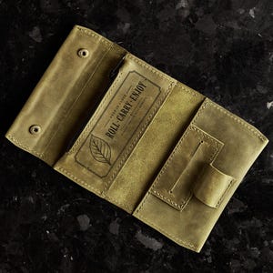 May include: An olive green leather tobacco pouch featuring a snap button closure, a zippered compartment, and a rectangular pocket. The interior is stamped with a leaf icon and the text "KEEP IT CLOSE ROLL CARRY ENJOY" in a decorative frame.