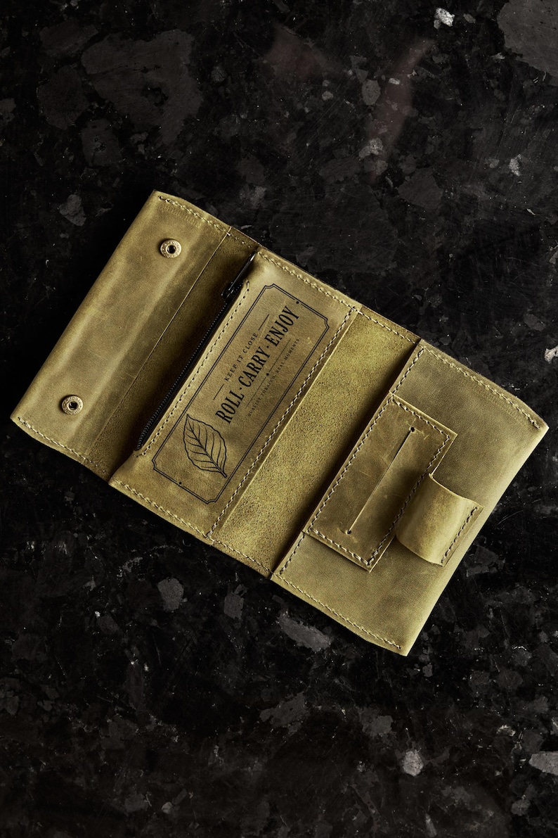 May include: An olive green leather tobacco pouch featuring a snap button closure, a zippered compartment, and a rectangular pocket. The interior is stamped with a leaf icon and the text "KEEP IT CLOSE ROLL CARRY ENJOY" in a decorative frame.