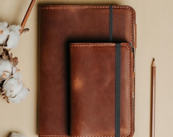 Peut inclure: Deux carnets en cuir marron, un grand et un petit, sont empilés. Chaque carnet a une bande élastique noire et un porte-stylo. Un crayon en bois et des brins de coton sont également dans l'image.