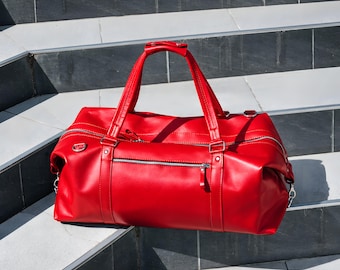 Könnte beinhalten: Eine leuchtend rote Reisetasche mit glatter Oberfläche, zwei Tragegriffen und einer horizontalen Reißverschlusstasche auf der Vorderseite. Die Tasche verfügt über silberfarbene Metallbeschläge, einschließlich Reißverschlüssen und seitlichen Ringen.