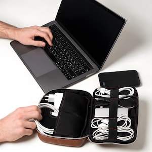Op de afbeelding: Een donkergrijze laptop met een zwart toetsenbord en een bruine, lederachtige organizer-etui met ritssluiting, gevuld met witte oplaadkabels, een witte stroomadapter en een zilveren multiport-hub. Een zwarte powerbank ligt ernaast.