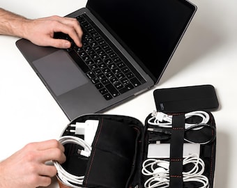 May include: A dark gray laptop with a black keyboard and a brown leather-like zippered organizer case containing white charging cables, a white power adapter, and a silver multi-port hub. A black rectangular power bank sits nearby.