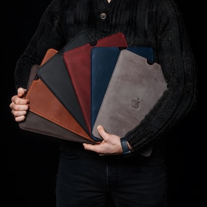 Könnte beinhalten: Eine Sammlung von sechs Laptop-Hüllen aus Leder in verschiedenen Farben, darunter Grau, Blau, Rot, Schwarz und Braun. Jede Hülle hat eine glatte Oberfläche und eine Aussparung an der Oberkante. Die graue Hülle zeigt ein eingeprägtes Apple-Logo.