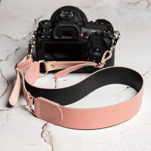 Può includere: Una tracolla per fotocamera rosa e nera con una fotocamera nera sullo sfondo. Il cinturino è realizzato in pelle e ha un lato inferiore nero imbottito per il comfort. La pelle rosa è cucita con filo bianco.