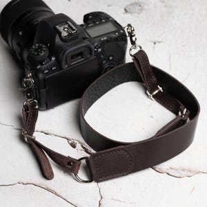 Op de afbeelding: Een zwarte DSLR-camera met een bruinleren camerariem. De riem is met metalen clips en ringen aan de camera bevestigd. De leren riem is donkerbruin en heeft een getextureerde onderkant.