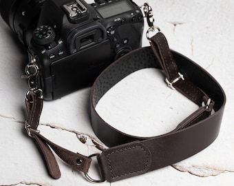 May include: A black DSLR camera with a dark brown leather camera strap. The strap is secured to the camera with metal clips and rings. The leather strap is a rich brown colour and has a textured underside.