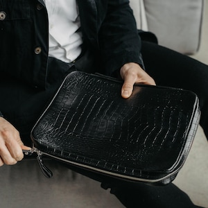 May include: A black rectangular zippered case featuring a textured crocodile skin pattern. The case has a silver-toned metal zipper that runs along the perimeter, with a matching black leather pull tab attached to the zipper slider for easy opening.