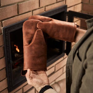 Può includere: Guanti da forno in pelle marrone con un anello per appenderli. I guanti sono tenuti davanti a un camino in mattoni con una porta in metallo nero. Proteggono le mani dal calore.