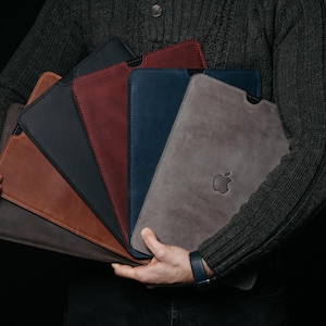 Può includere: Una collezione di custodie per tablet in pelle in vari colori, tra cui marrone, blu navy e grigio. Ogni custodia presenta un design elegante e un logo Apple in rilievo. Le custodie sono disposte a ventaglio.