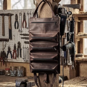 May include: A dark brown leather tool roll featuring five horizontal zippered compartments and a top handle for hanging. The organiser has a rectangular shape with visible stitching along the edges and a bottom strap with a metal buckle closure.
