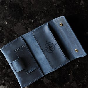 May include: A blue leather wallet featuring a tri-fold design with visible stitching, a small pen loop, and a central compass rose engraving. The interior includes multiple card slots and two brass-colored metal snap fasteners for closure.