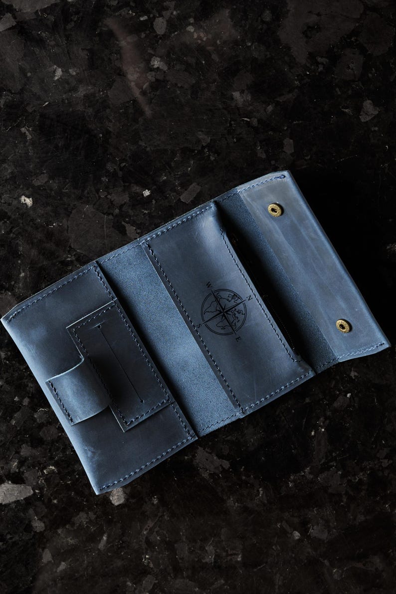 May include: A blue leather wallet featuring a tri-fold design with visible stitching, a small pen loop, and a central compass rose engraving. The interior includes multiple card slots and two brass-colored metal snap fasteners for closure.