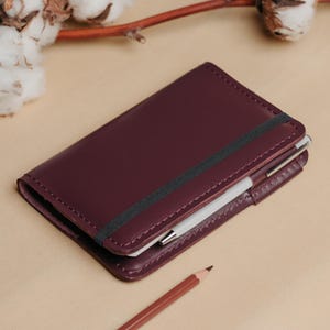 May include: A deep burgundy leather notebook cover featuring thick, visible stitching along the edges. A black elastic band wraps around the centre to keep the cover closed, and a white pen is tucked into a side loop on the right edge.