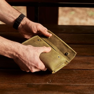 May include: An olive green leather wallet featuring a fold-over design with visible stitching along the edges and three metallic snap button closures. The wallet has a matte finish and a rectangular shape, showing the front flap partially open.