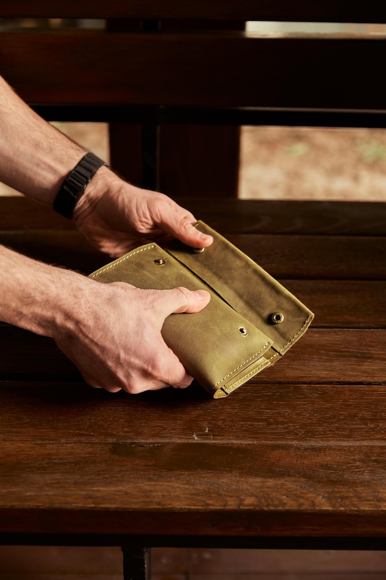 May include: An olive green leather wallet featuring a fold-over design with visible stitching along the edges and three metallic snap button closures. The wallet has a matte finish and a rectangular shape, showing the front flap partially open.