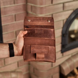 May include: A brown leather tobacco pouch featuring a fold-over flap with two metallic snap buttons, an internal zippered compartment, and a rectangular pocket with a central strap closure, showcasing visible stitching along the edges.