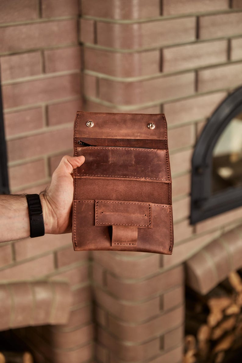 May include: A brown leather tobacco pouch featuring a fold-over flap with two metallic snap buttons, an internal zippered compartment, and a rectangular pocket with a central strap closure, showcasing visible stitching along the edges.