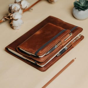 May include: Two brown leather-covered notebooks stacked on top of each other, each featuring a dark elastic band closure. A metallic pen is tucked into the elastic of the smaller notebook, and a second pen rests between the two covers.