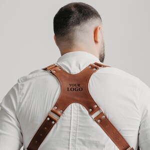 Könnte beinhalten: Brauner Leder-Kameragurt mit silberfarbenen Beschlägen. Der Gurt hat ein Kreuz-Design auf dem Rücken und der Text "YOUR LOGO" ist auf der Rückseite eingeprägt. Getragen über einem weißen Hemd.