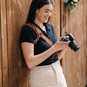 May include: A brown leather camera strap with metal fixings is worn over a dark blue shirt. The camera strap is attached to a black camera with a lens. The person is wearing beige trousers and is leaning against a wooden wall.