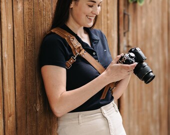 May include: A brown leather camera strap with metal fixings is worn over a dark blue shirt. The camera strap is attached to a black camera with a lens. The person is wearing beige trousers and is leaning against a wooden wall.