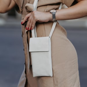 May include: A light gray, textured leather crossbody phone bag with a flap closure. The bag has a long, thin strap. The person is wearing a tan dress and a silver watch.
