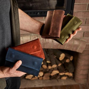 May include: Four rectangular leather wallets in blue, orange, brown, and olive green. Each wallet features a fold-over flap with a single snap button closure and visible stitching along the edges, showcasing a matte finish and natural texture.