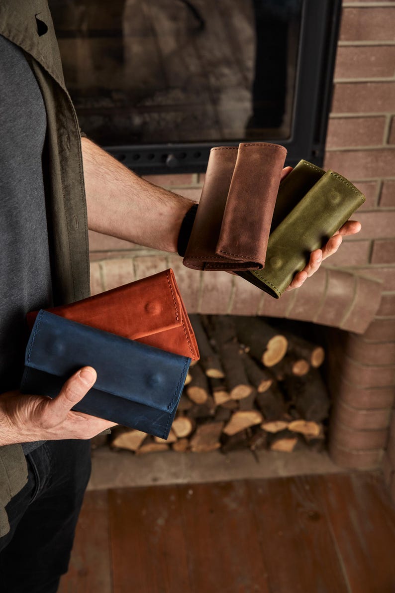May include: Four rectangular leather wallets in blue, orange, brown, and olive green. Each wallet features a fold-over flap with a single snap button closure and visible stitching along the edges, showcasing a matte finish and natural texture.